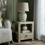 Side table beside a cream armchair, styled with a green floral lamp on top and a small cream vase on books on the open shelf.