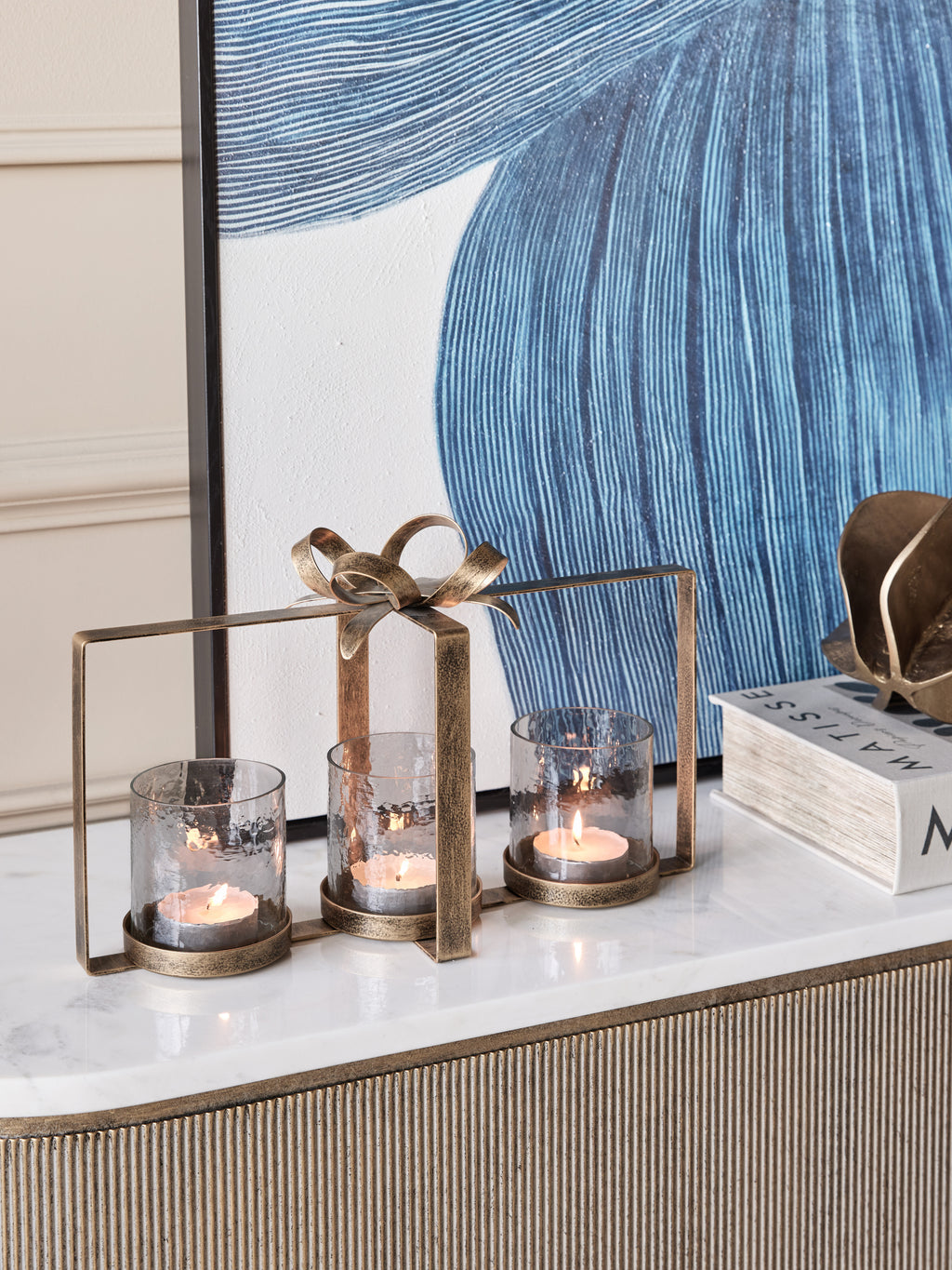 Three lit candles in the bow hurricane on a pale sideboard top, with blue artwork and books behind.