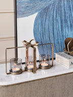 Three lit candles in the bow hurricane on a pale sideboard top, with blue artwork and books behind.