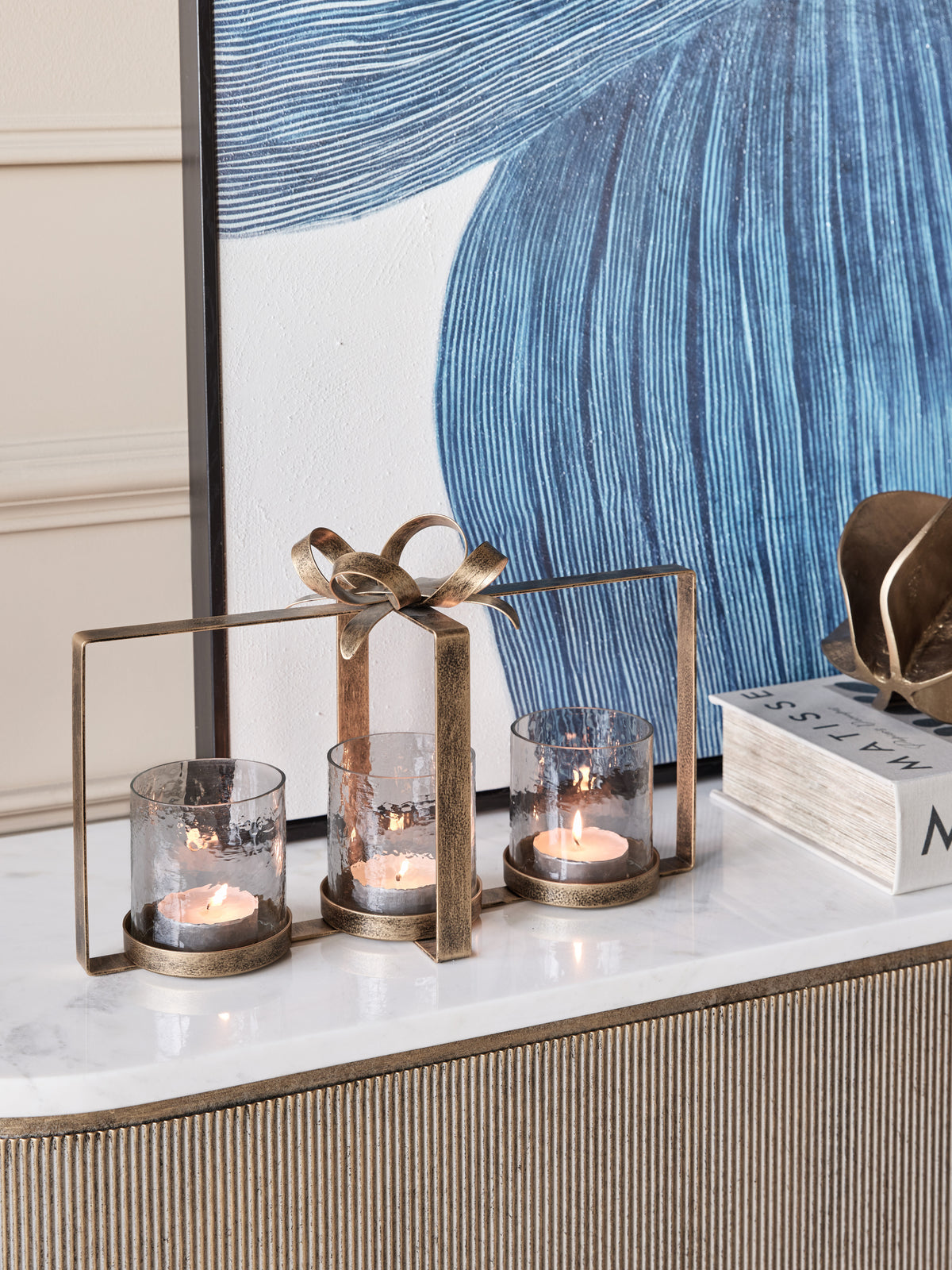Three lit candles in the bow hurricane on a pale sideboard top, with blue artwork and books behind.
