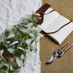 Artificial sage-green foliage bouquet resting on white cloth beside an open blank notebook and pruning shears on a tan fabric background.