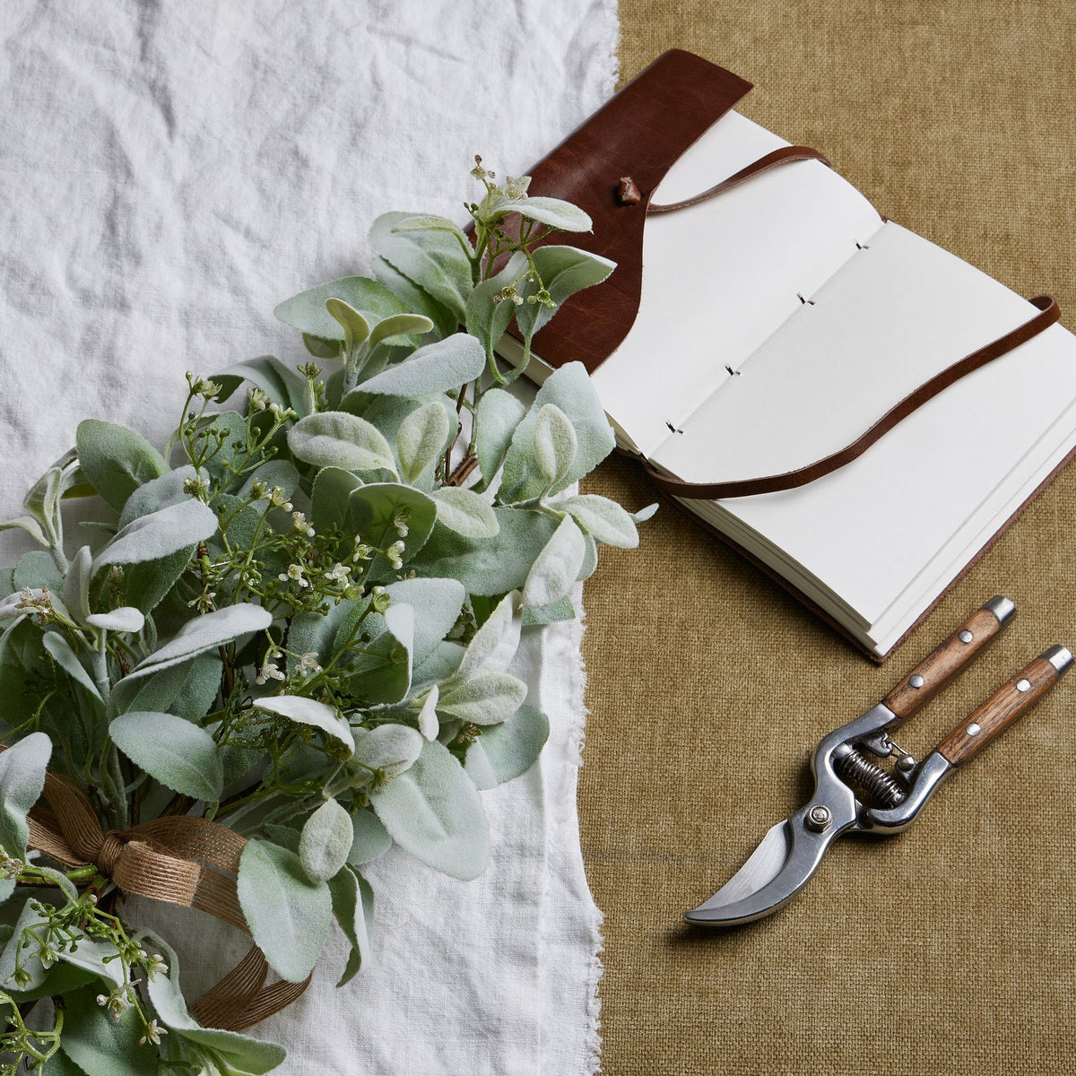 Artificial sage-green foliage bouquet resting on white cloth beside an open blank notebook and pruning shears on a tan fabric background.