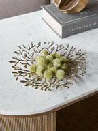 Leaf platter filled with green decorative seed heads on a white veined tabletop beside a book and gold ornament.