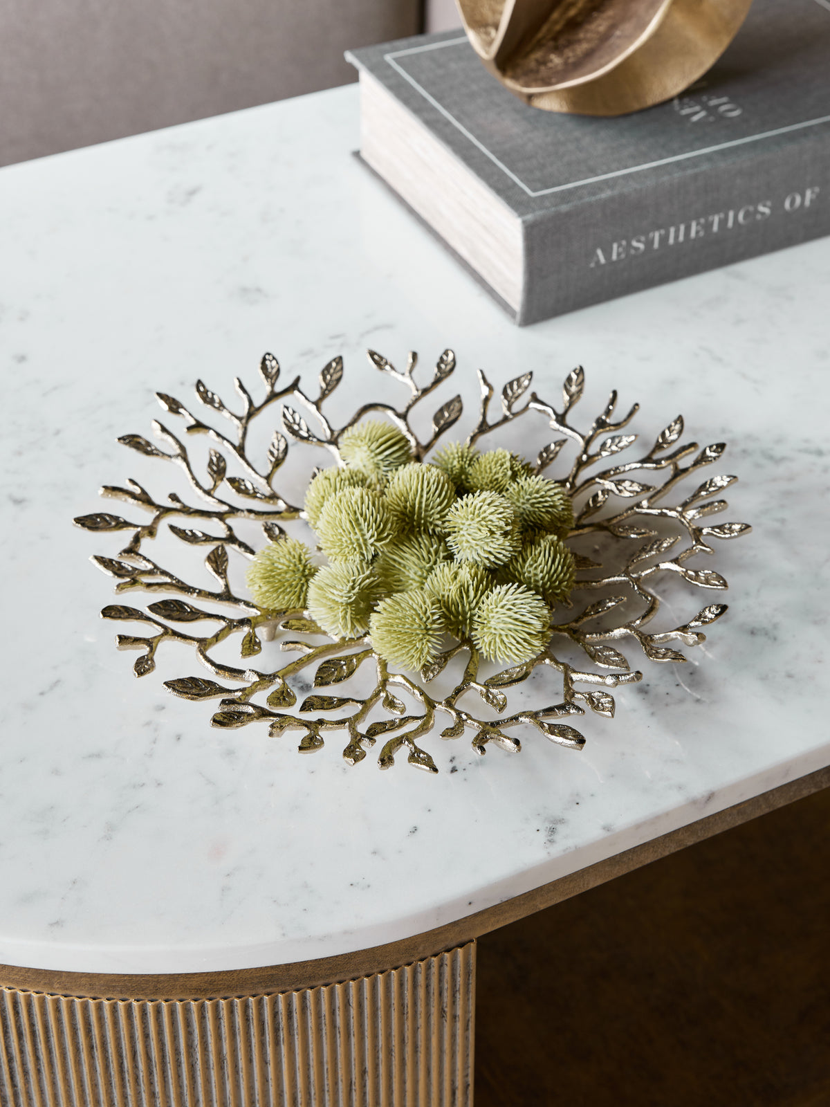 Leaf platter filled with green decorative seed heads on a white veined tabletop beside a book and gold ornament.