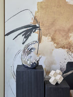 Silver wave sculpture on a dark ribbed pedestal in front of abstract artwork, beside a pale geometric ornament on a lower pedestal.