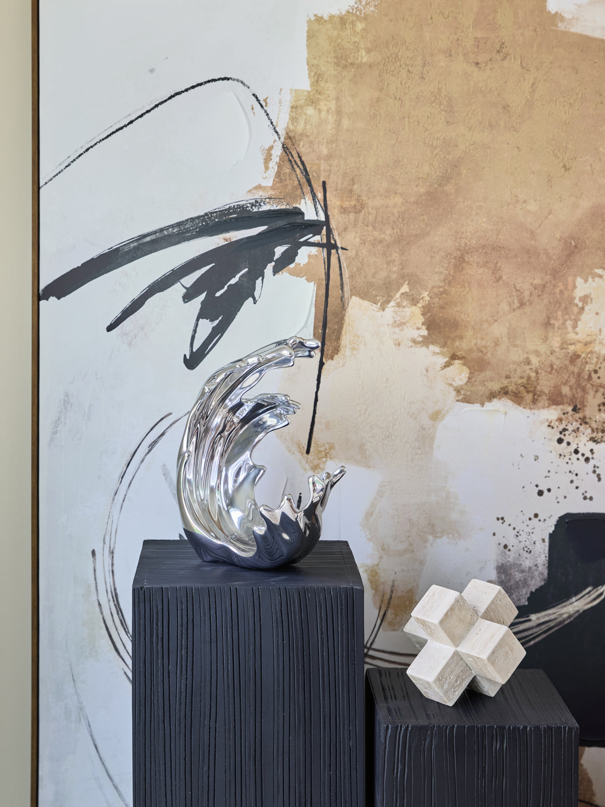 Silver wave sculpture on a dark ribbed pedestal in front of abstract artwork, beside a pale geometric ornament on a lower pedestal.