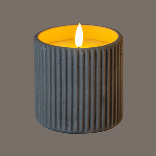 Ribbed cylindrical LED candle photographed illuminated, with a bright flame-shaped centre and amber light inside the rim; the outer wall appears dark grey against a taupe background.