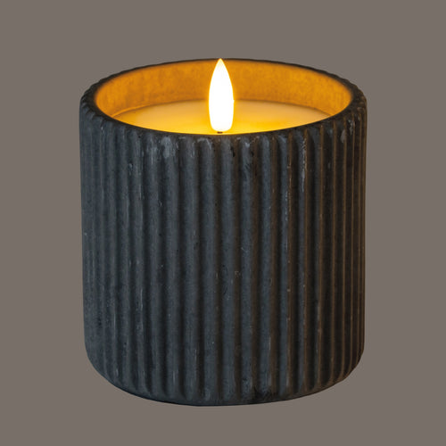 Ribbed LED candle shown illuminated, with a bright flame-shaped element and amber light around the recessed top; the grey wall appears dark in this lighting.