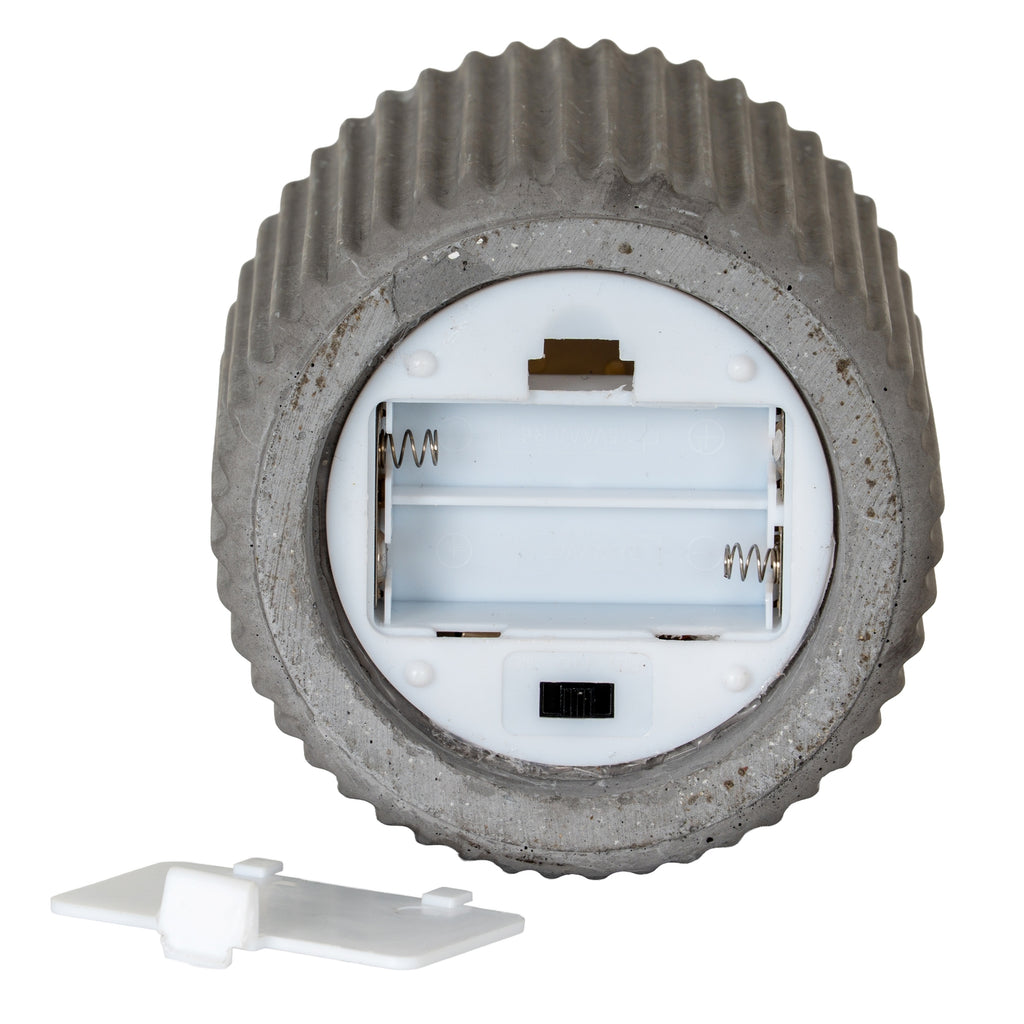 Underside of the grey ribbed candle showing a round white insert, open empty two-channel battery compartment, metal-looking contacts and black switch, with the white cover detached below.