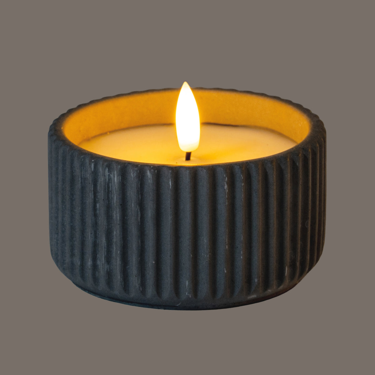 Ribbed LED candle photographed illuminated, with a bright flame-shaped centre and amber light across the inset top; the grey wall appears dark in this lighting.