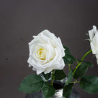 Close view of a layered white artificial rose with a cream centre, veined green leaves and part of a second flower at the right.