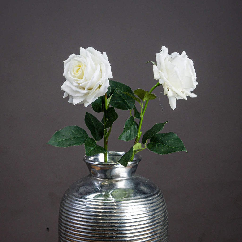 Two white artificial garden roses with dark green leaves arranged in a ribbed silver-coloured vase against a grey background.