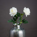 Two white artificial garden roses with dark green leaves arranged in a ribbed silver-coloured vase against a grey background.