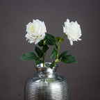 Two white artificial garden roses with dark green leaves arranged in a ribbed silver-coloured vase against a grey background.