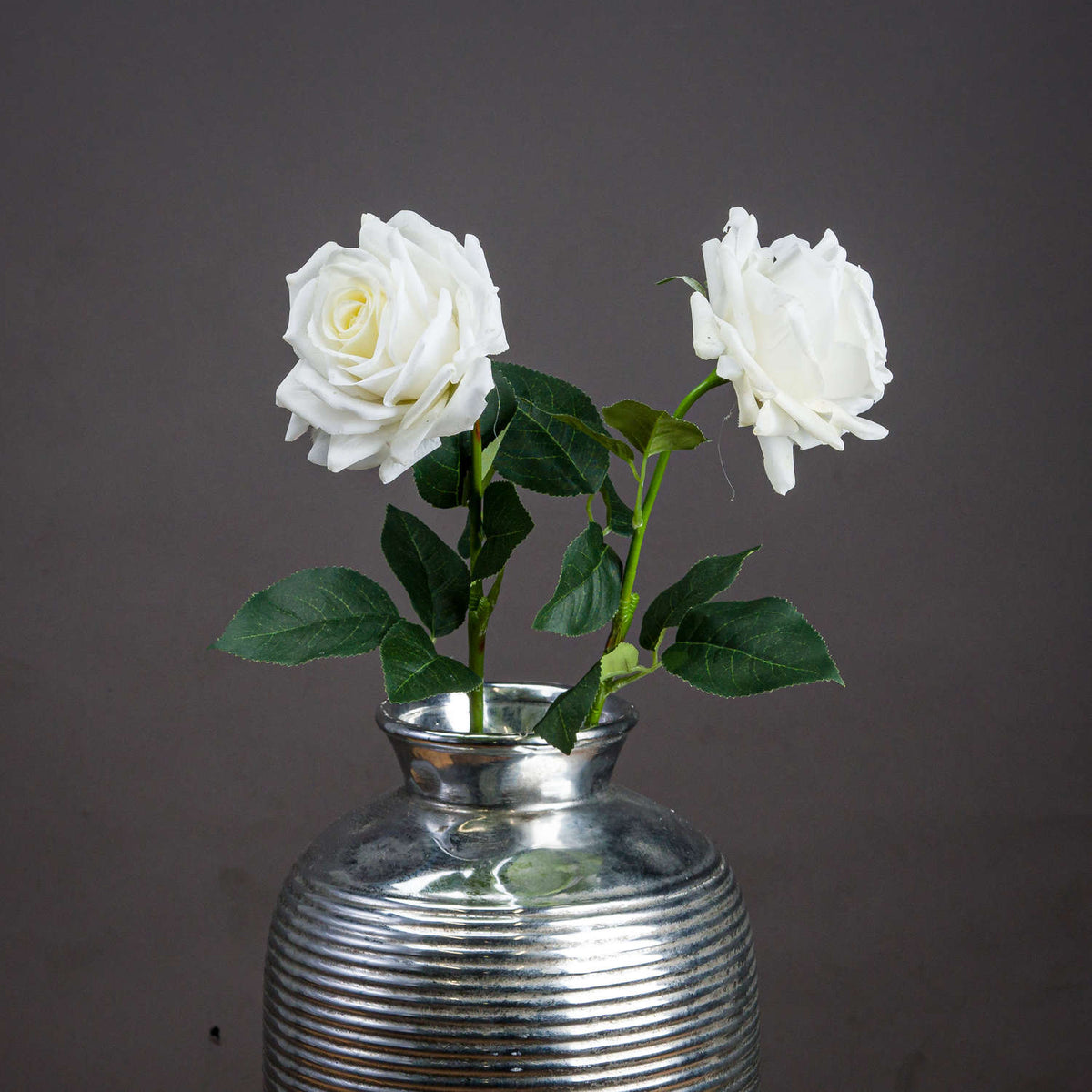 Two white artificial garden roses with dark green leaves arranged in a ribbed silver-coloured vase against a grey background.