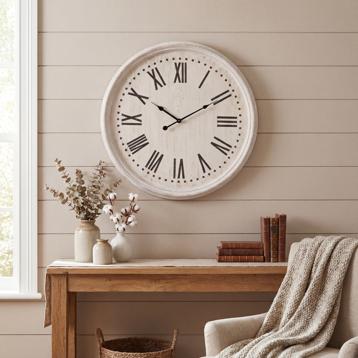 Pale Roman numeral wall clock on a taupe panelled wall above a wooden console with ceramic vases and books, beside a cream armchair.