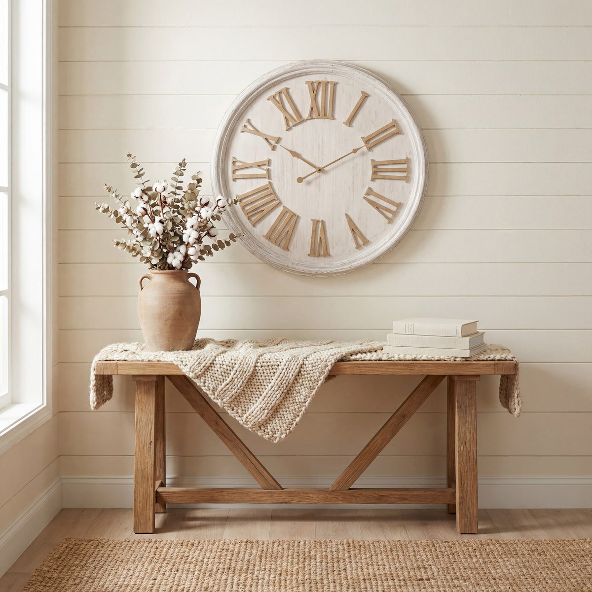 Whitewashed Roman numeral wall clock on a cream panelled wall above a wooden console styled with a knitted throw, books and a vase of stems.