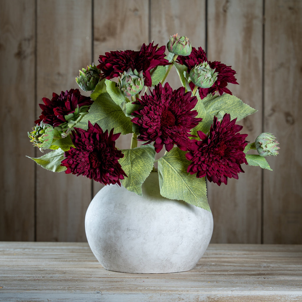 Several burgundy artificial flower stems arranged with green buds and leaves in a rounded pale vase on a wooden surface.