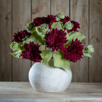 Several burgundy artificial flower stems arranged with green buds and leaves in a rounded pale vase on a wooden surface.