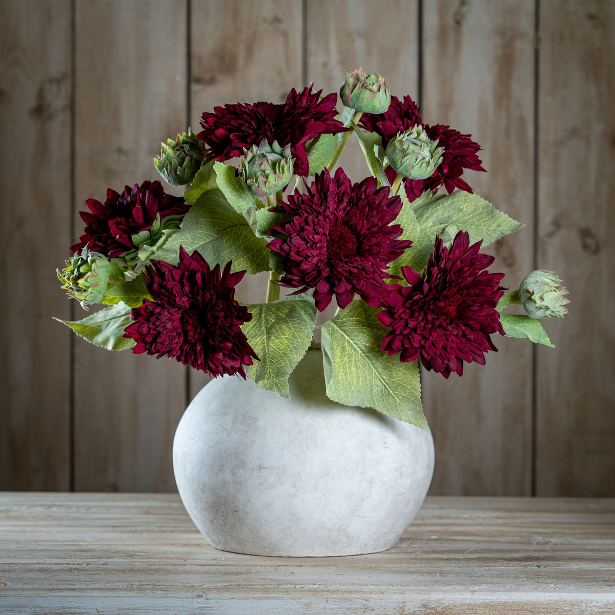 Several burgundy artificial flower stems arranged with green buds and leaves in a rounded pale vase on a wooden surface.