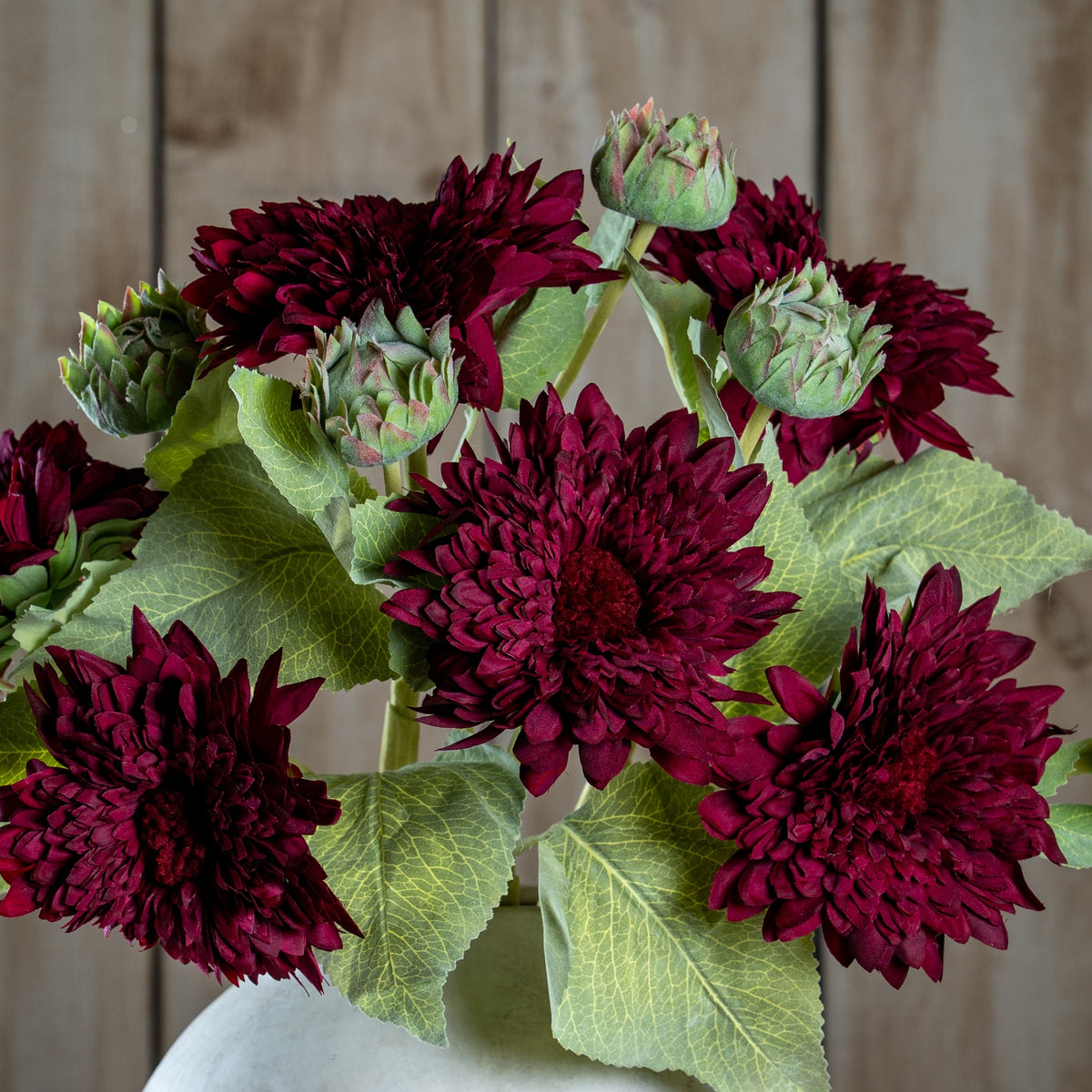 Close-up of a grouped burgundy floral arrangement showing layered blooms, green buds and veined leaves against wooden panelling.