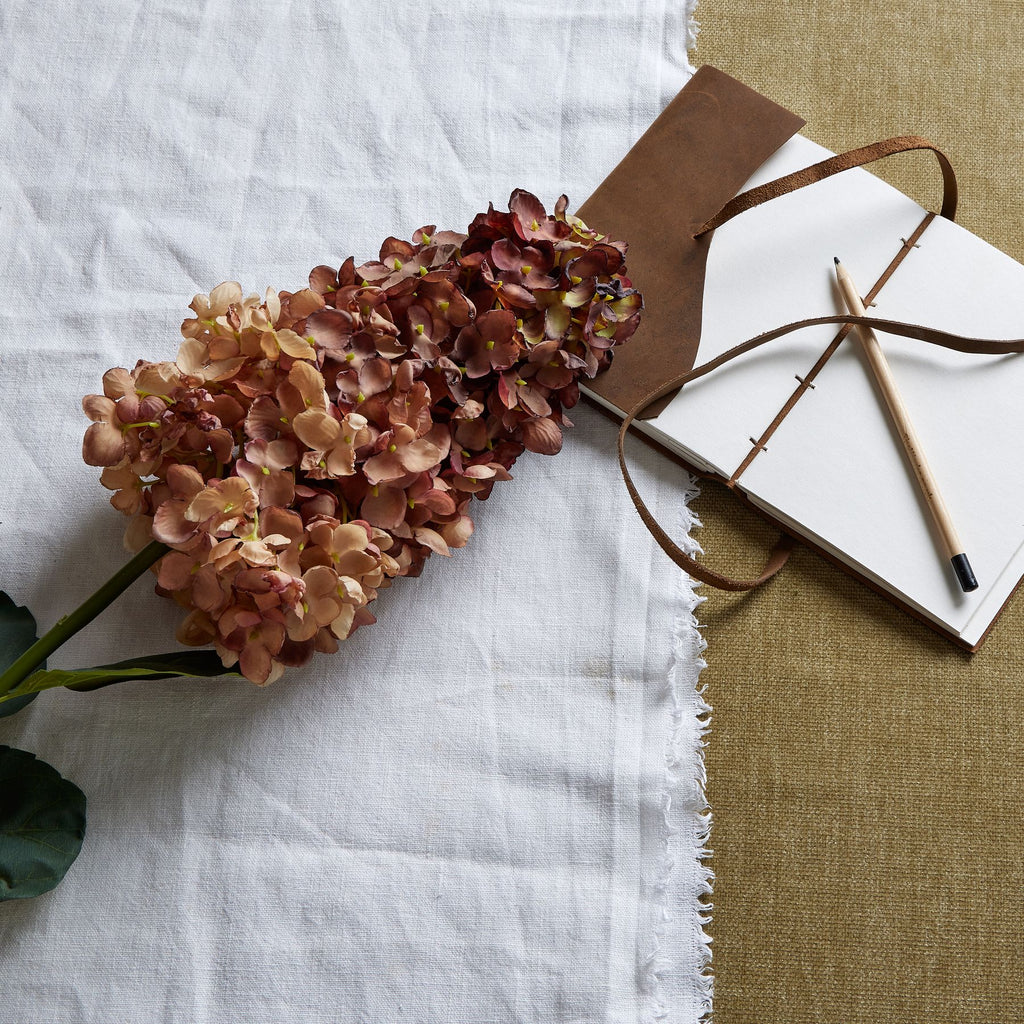 Burgundy and tan artificial flower head laid diagonally on white fabric beside an open brown-covered notebook and a pencil.