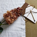 Burgundy and tan artificial flower head laid diagonally on white fabric beside an open brown-covered notebook and a pencil.