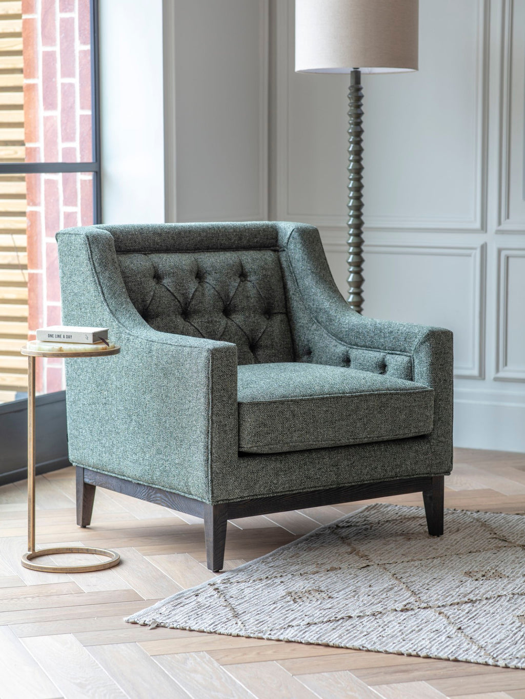 Green buttoned armchair beside a small round side table and floor lamp near a window, with a pale textured rug.