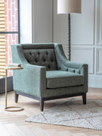 Green buttoned armchair beside a small round side table and floor lamp near a window, with a pale textured rug.