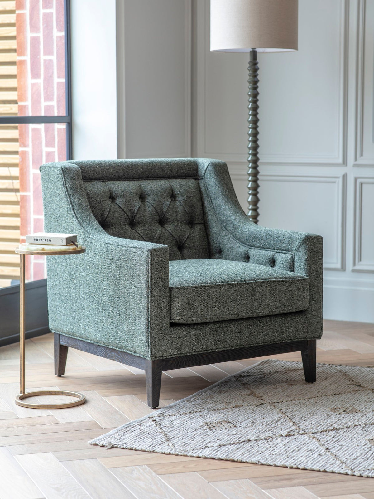 Green buttoned armchair beside a small round side table and floor lamp near a window, with a pale textured rug.