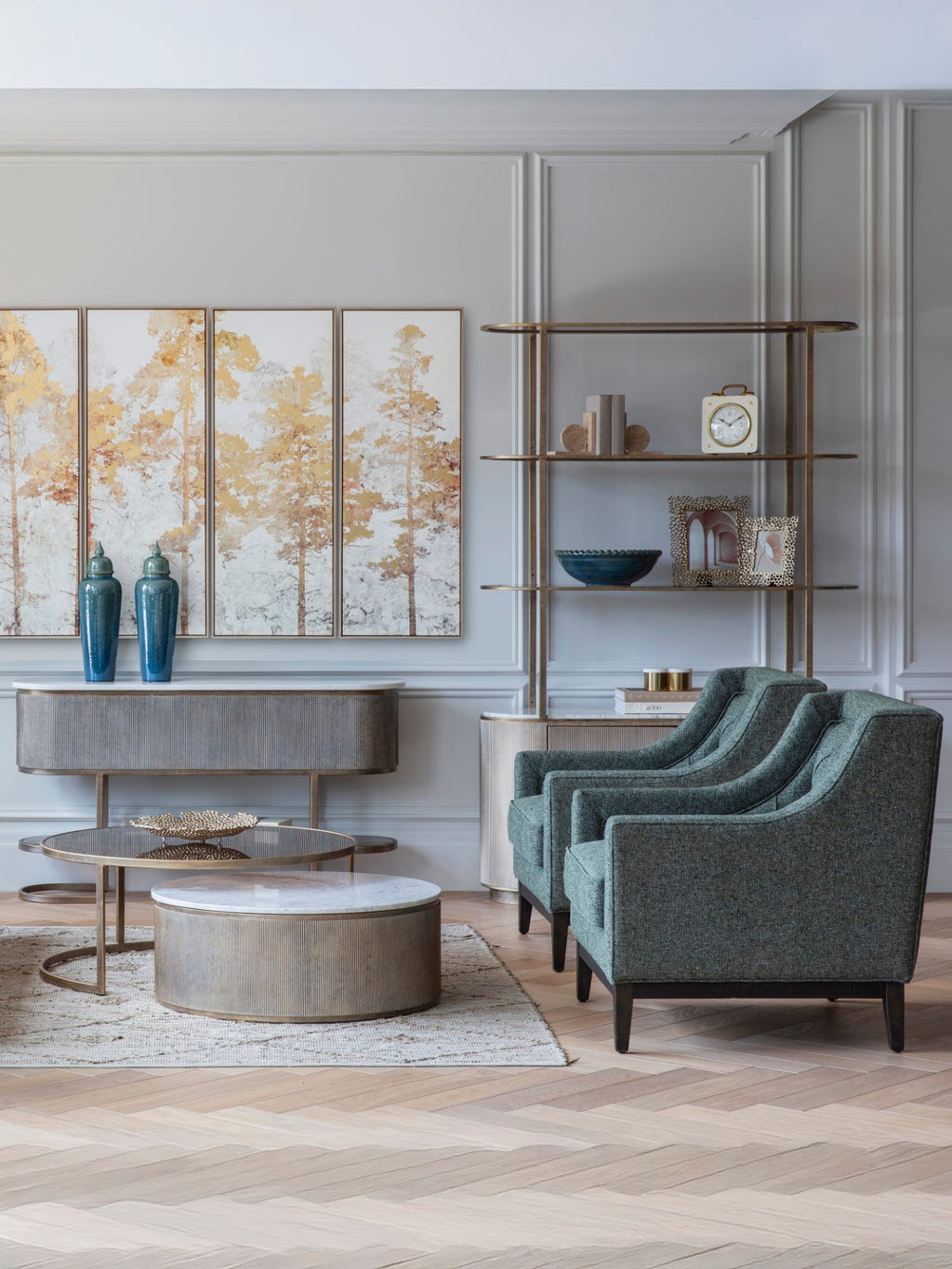 Two green armchairs beside round coffee tables in a panelled living room with gold-toned shelving and tree artwork.