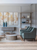 Two green armchairs beside round coffee tables in a panelled living room with gold-toned shelving and tree artwork.