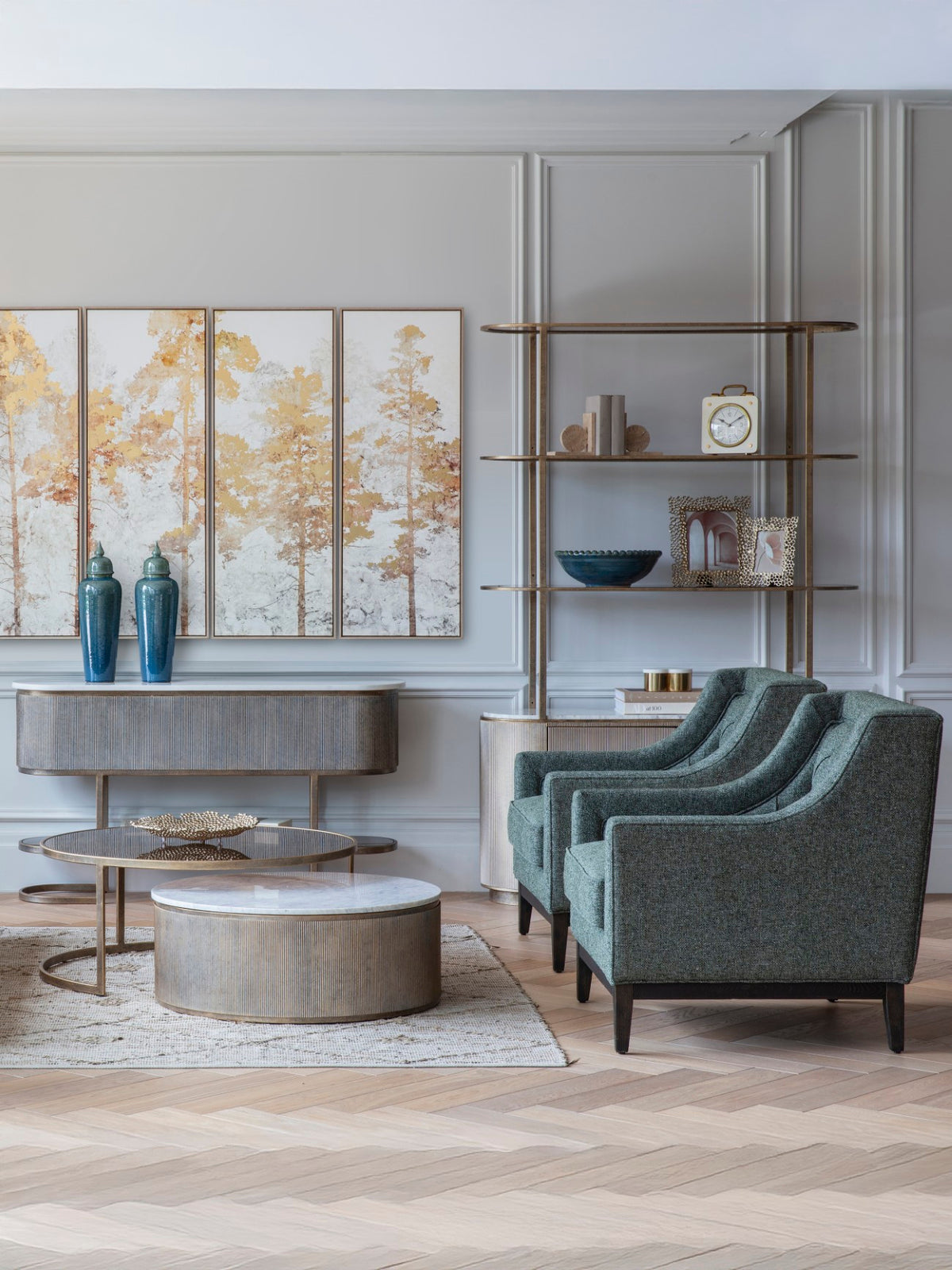 Two green armchairs beside round coffee tables in a panelled living room with gold-toned shelving and tree artwork.
