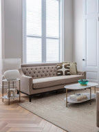 Taupe buttoned sofa styled with patterned cushions beside a lamp and oval coffee table in a pale room with shuttered windows.