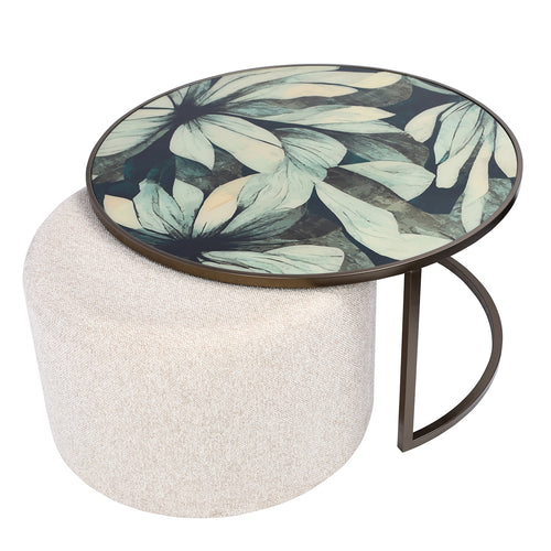 Elevated angled view of the pale leaf-patterned tabletop, with the round oatmeal pouffe extending beneath it to the left.