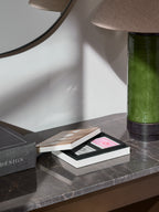 Open card box with black-backed and red-backed card packs on a dark veined console beside a green lamp and book.