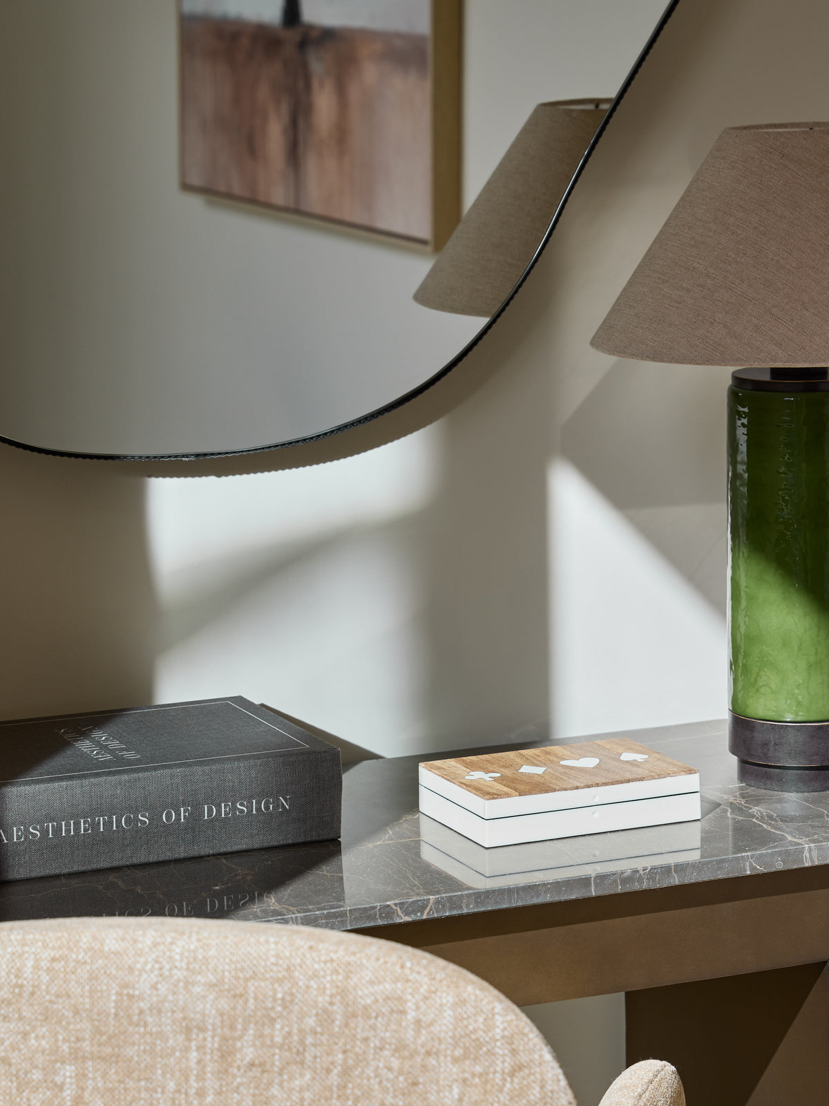 Closed card box on a dark veined console between a grey book and green lamp, beneath a round mirror.