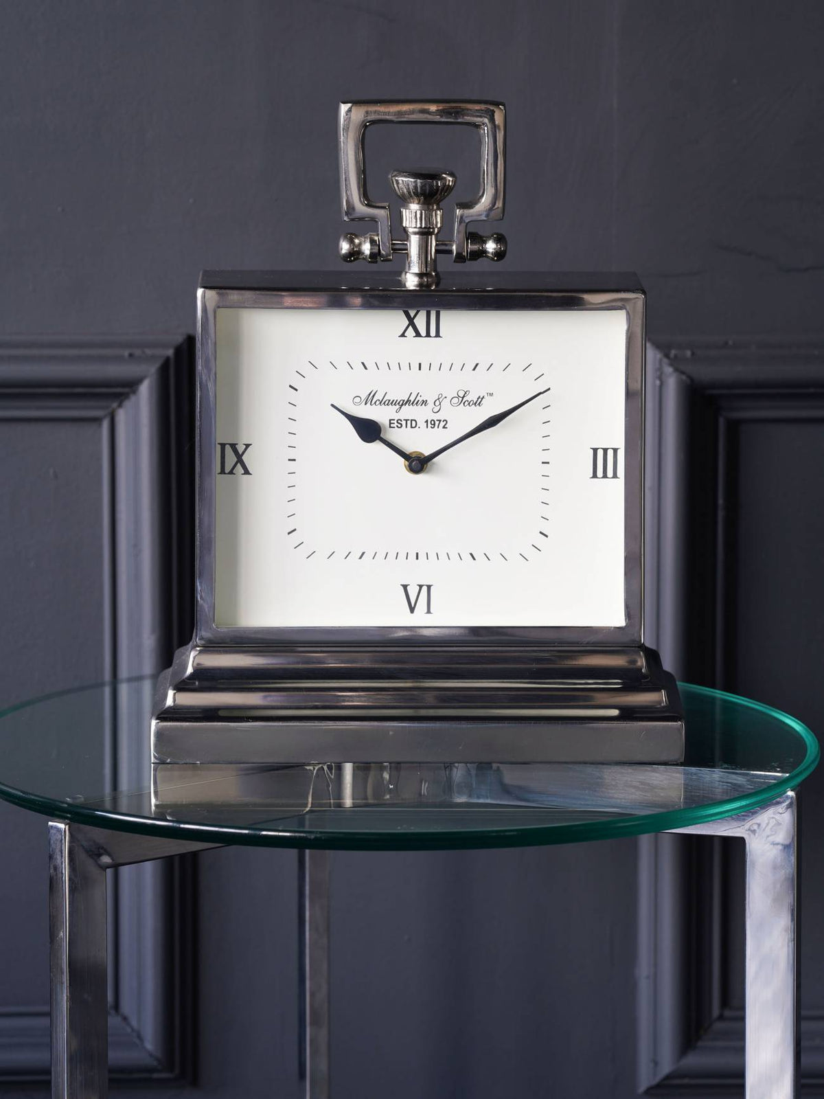 Polished silver-coloured carriage clock standing on a round glass table against a dark panelled wall.
