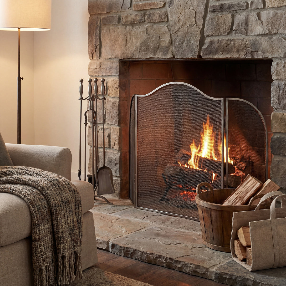 Arched silver-tone fire screen pictured before a lit stone fireplace, with fireside tools, a log basket, floor lamp and upholstered armchair in the surrounding scene.