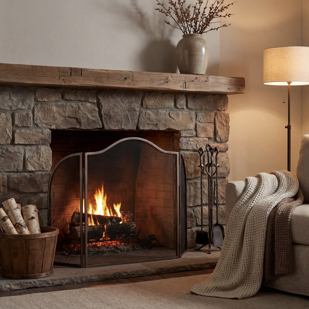 Silver-tone arched fire screen styled in front of a lit stone fireplace with a timber mantel, log basket, fireside tools and an armchair with a knitted throw.