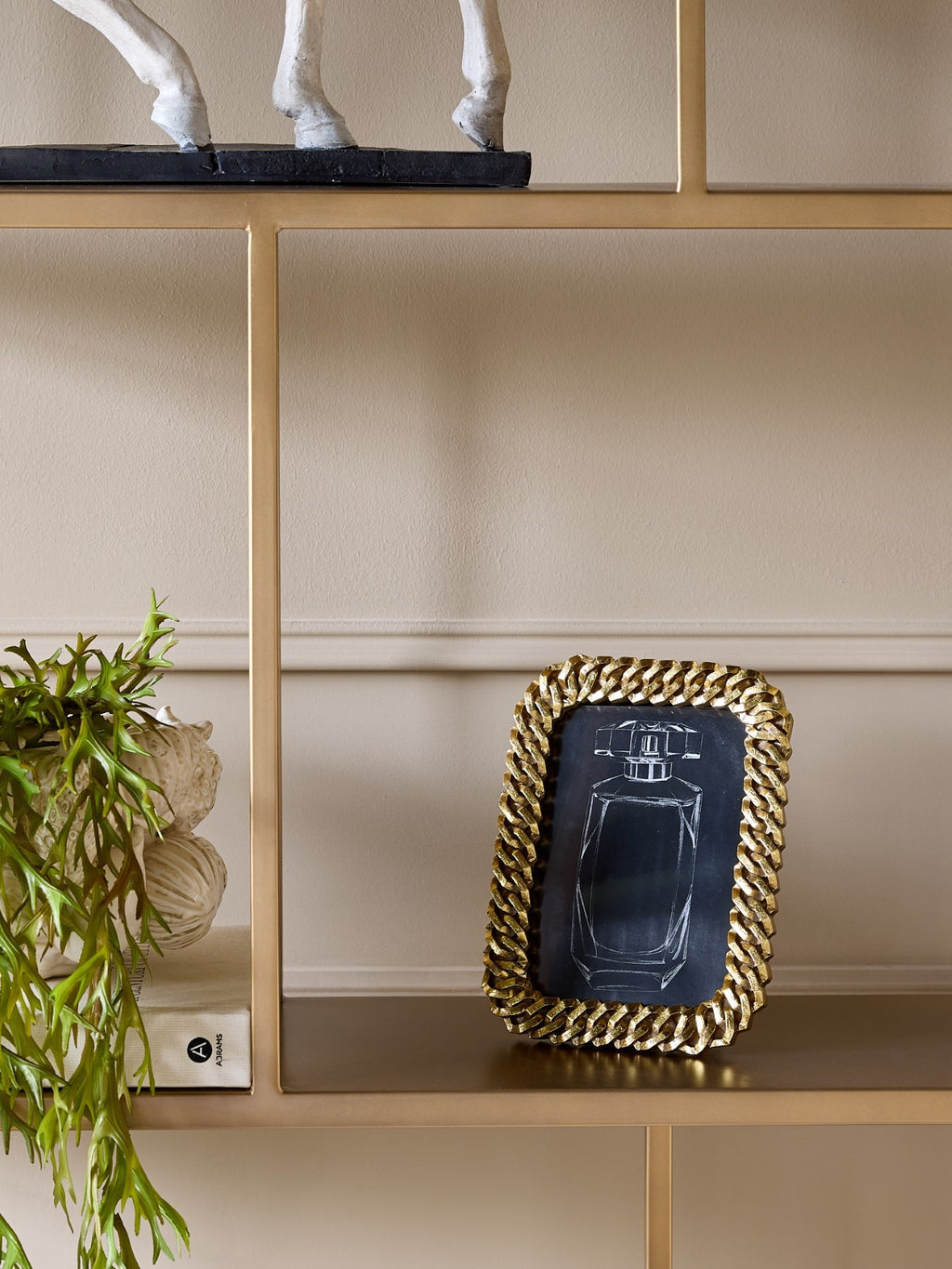 Gold chain-link photo frame standing on a gold shelving unit beside books and a trailing green plant.