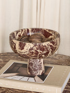 Red marble footed bowl filled with brown decorative balls, standing on a large book on a patterned tabletop.