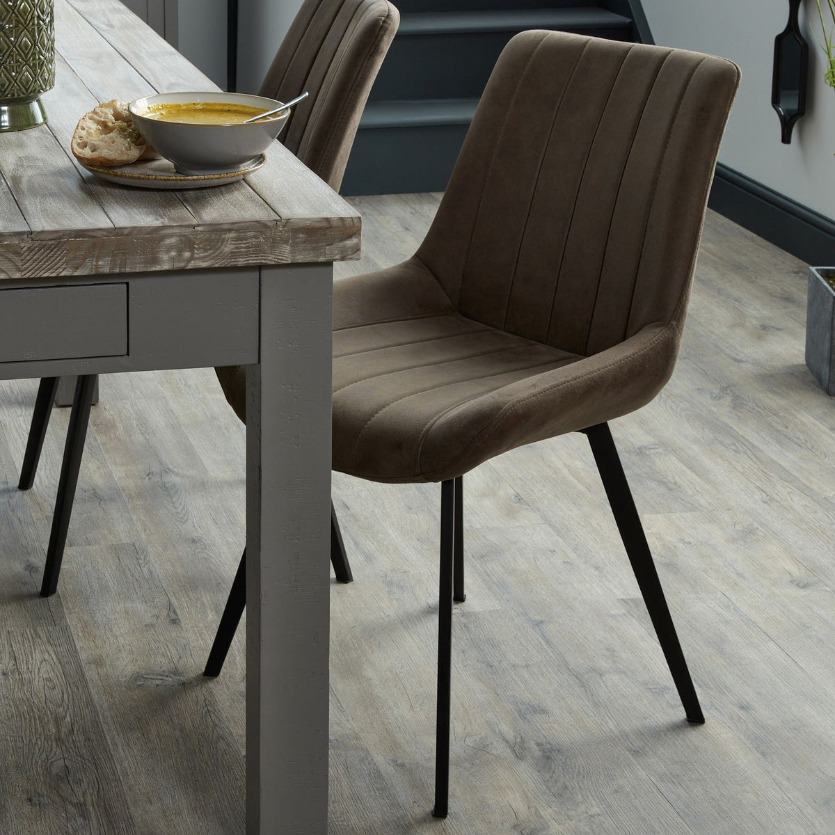 Brown channelled dining chair beside a grey wooden dining table set with soup and bread, with a matching chair behind.