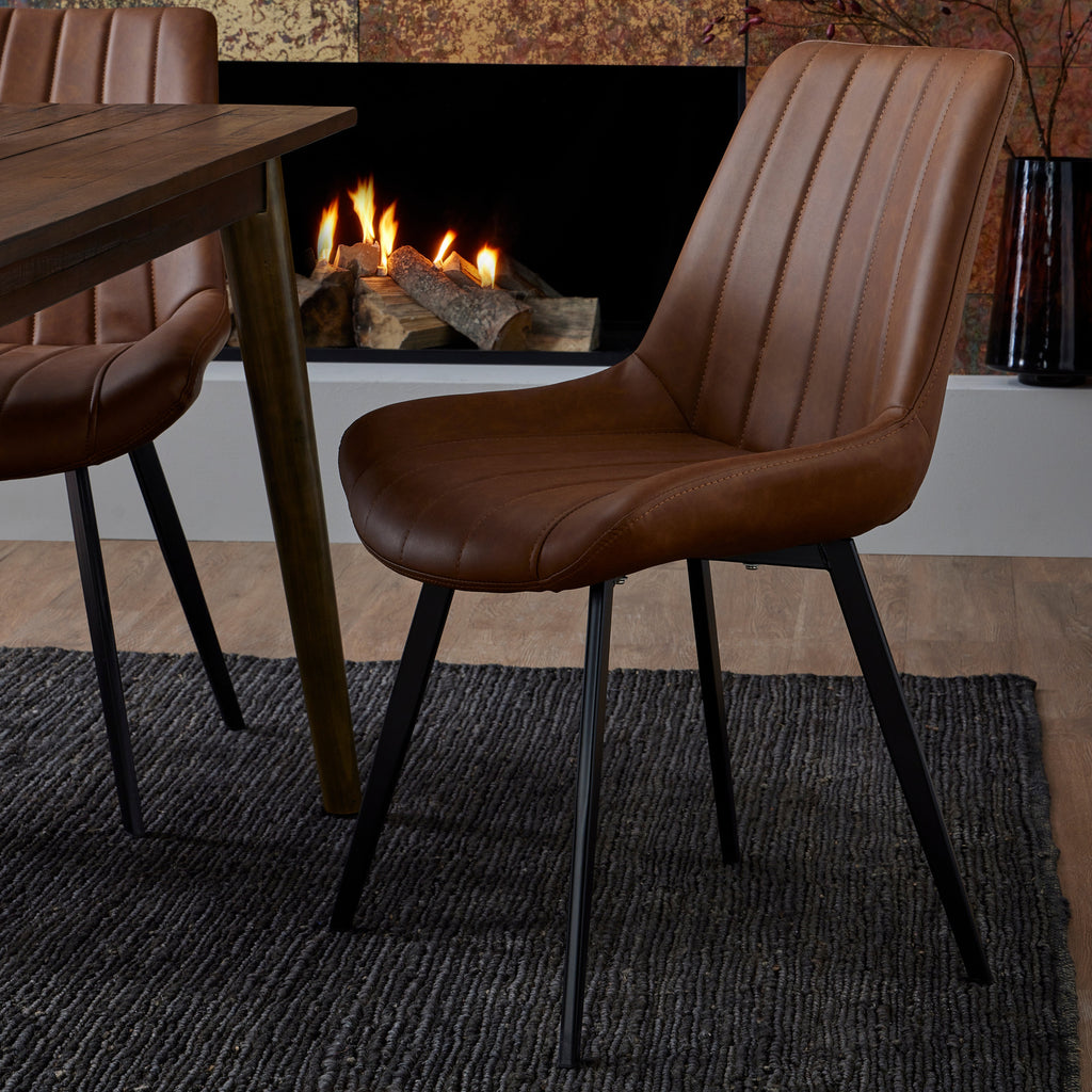 Tan channel-seamed dining chair beside a dark timber table on a charcoal rug, with part of a second chair, a lit fireplace and a black vase in the background.