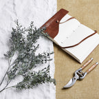 Charcoal foliage branches arranged diagonally on white cloth beside an open brown-covered notebook and pruning shears on a tan woven surface.