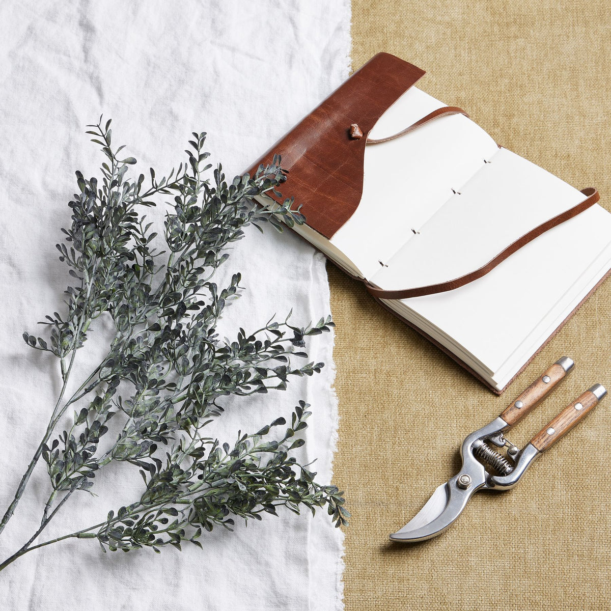 Charcoal foliage branches arranged diagonally on white cloth beside an open brown-covered notebook and pruning shears on a tan woven surface.