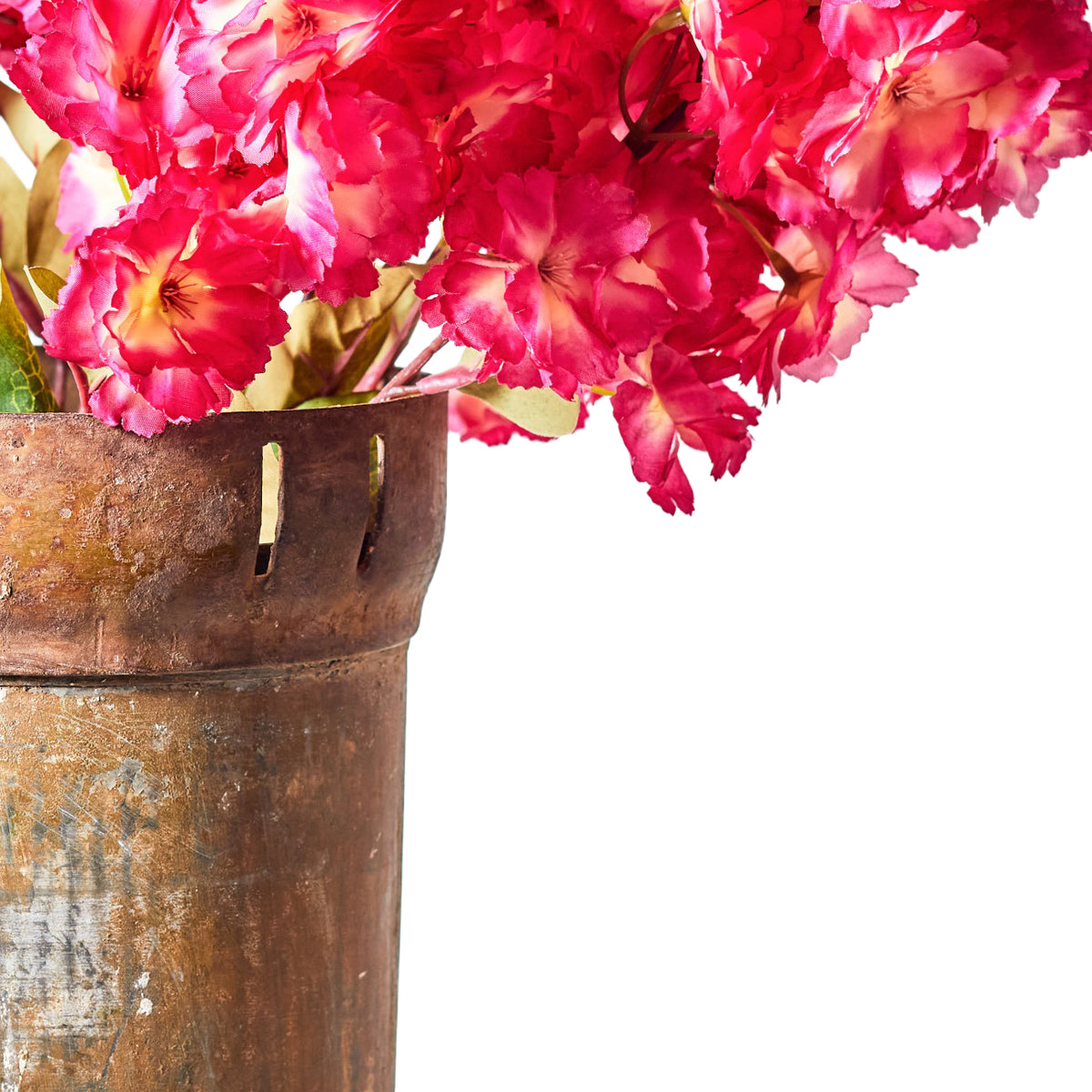Close-up of clustered pink and cherry-red faux blossoms with pale centres, dark stamens and green leaves in a weathered brown metallic vessel.