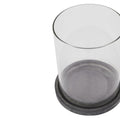Close angled view of an empty clear cylindrical lantern sleeve seated in a shallow dark grey round base with pale weathered markings, against white.