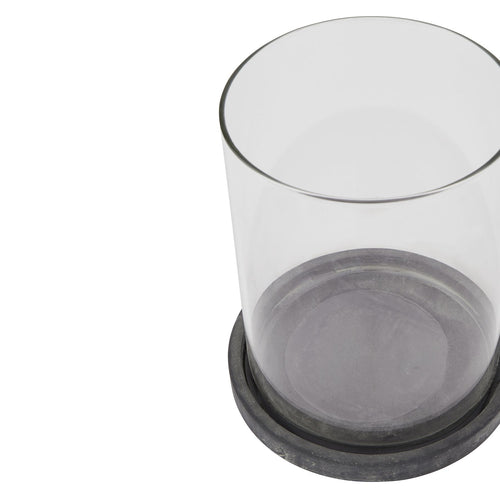 Close angled view of an empty clear cylindrical lantern sleeve seated in a shallow dark grey round base with pale weathered markings, against white.