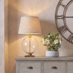 Table lamp pictured lit on a grey timber sideboard beside white flowers in a grey pot, beneath part of a large open-face wall clock.